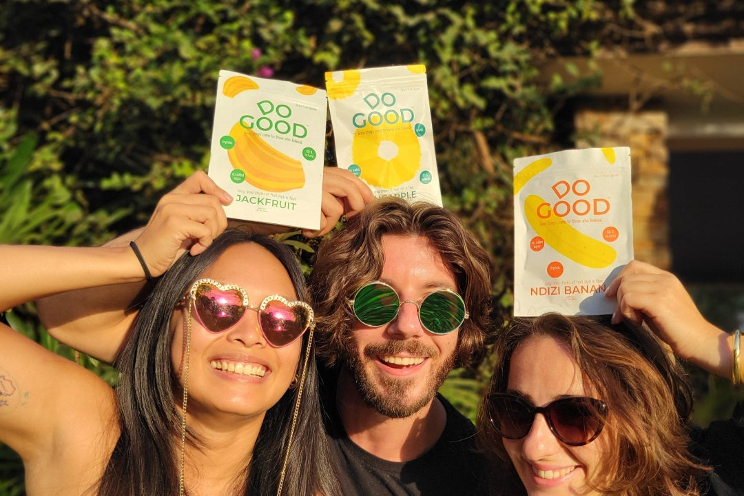 Group of friends smiling and holding DoGood organic dried fruit snack packs of pineapple, ndizi banana, and jackfruit outdoors in the sunshine