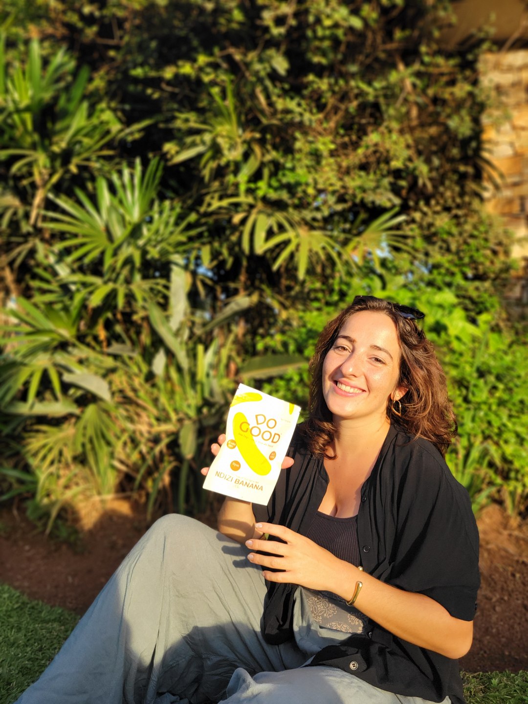 Smiling woman sitting outdoors holding a DoGood organic dried ndizi banana fruit snack pack in natural sunlight