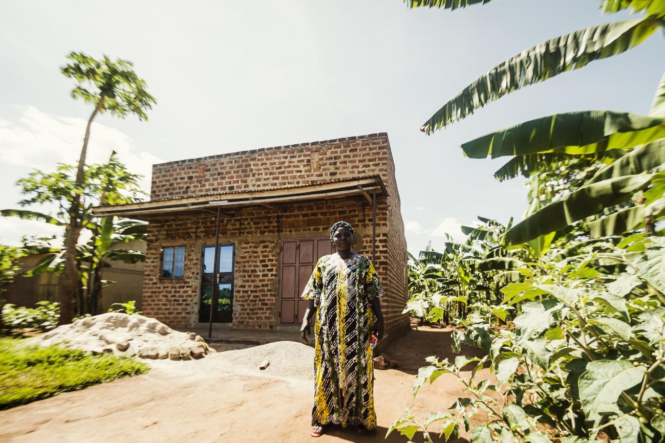 Ugandan woman farmer standing proudly in front of her brick home surrounded by banana trees – showcasing DoGood Earth’s commitment to sustainable farming communities